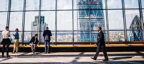 A large window view shows a view of London cityscape. Two people in suits and two people in casual clothing stand and sit near to the window. A man in a black suit walks along the tiled floor to the left.