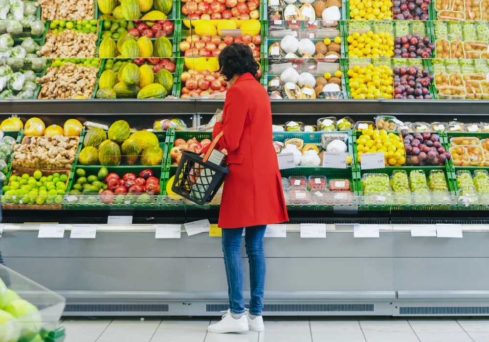 A woman in a red coat picks fruit from the shelves at a supermarket and puts it in her basket.