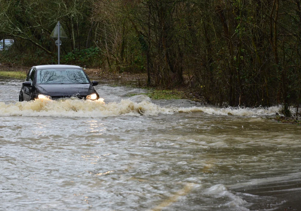 A car with headlights on struggles to drive through a waterlogged area.