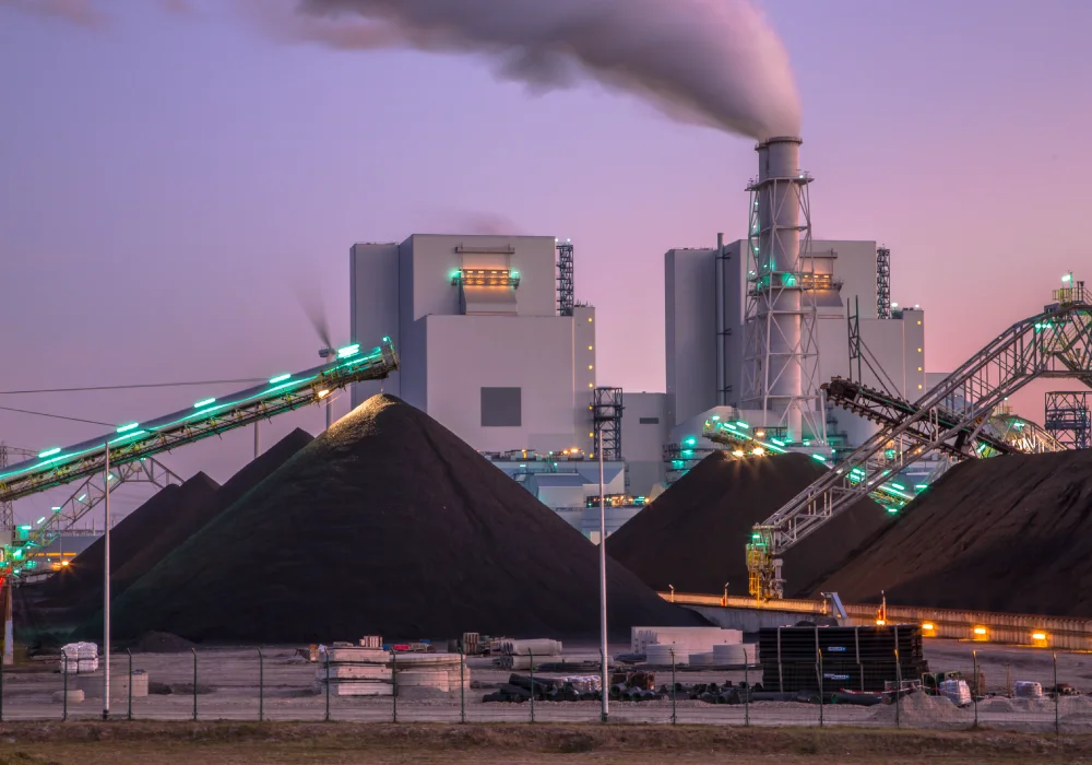 A large coal power station in purple sunset lighting. Large pyramids of coal can be seen, illuminated by the dwindling sun and bright lighting.