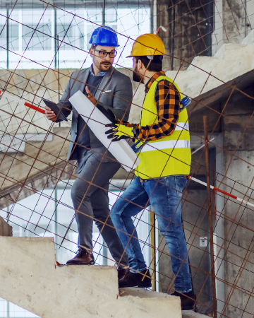 Two men in hard hats walk up some steps at a construction site. One man has a hi-vis vest and is carrying a large sheet of paper.